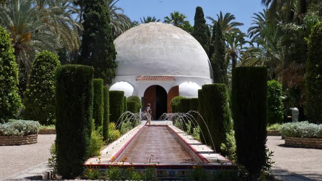 A white domed building in the Spanish city of Elche. On a water basin with fountains, a man refreshes himself and cools off with water. Sunshine and blue sky.