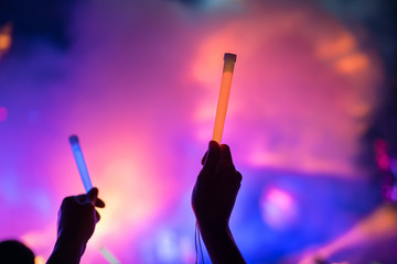 Young teenager girl fan raised up two hands with colored glowing sticks supporting favorite popular band on the night concert. Colorful crowdy background entertainment with light and laser show.