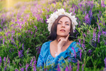 Young pretty woman with flower wreath sitting in purple field. Portrait of girl in blue clothing.