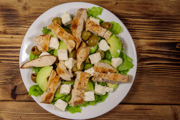 Tasty salad with fried chicken breast, green olives, feta cheese, avocado, lettuce and olive oil on wooden table. Top view