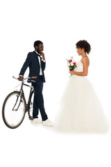 african american bridegroom standing near bicycle and looking at cheerful bride with flowers isolated on white