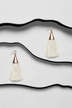 Shot of long earrings with pendants in the view of metal triangle with milken tassels. The accessory set is isolated asymmetrically against the white background between black faux leather stripes. 
