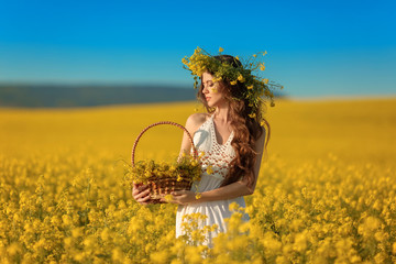 Beautiful young woman with wreath on long healthy hair over Yellow rape field landscape background. Attracive brunette girl with curly hairstyle holding basket with flowers, outdoor portrait.