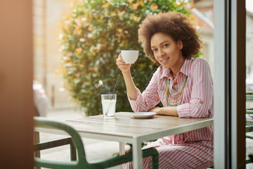 Beautiful smiling mixed race woman in pink striped dress sitting in cafe and enjoyingh her coffee.