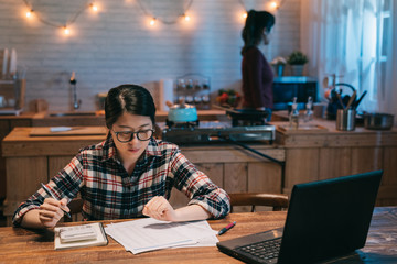 Attractive young housewife wearing shirt at home studying gas and electricity bills checking calculations looking at sheet of paper serious focused express night. roommates standing in home kitchen