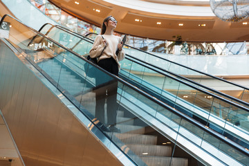 Full length photo of beautiful pretty woman smiling and holding clipboard while going down escalator in building indoors