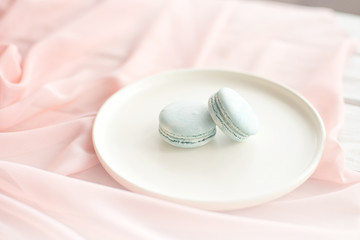 Obraz na płótnie Canvas Blue macaroni or macaroon cakes with pink textile flowers on a white ceramic plate on a gray wooden background. Flat lay, top view, copy space