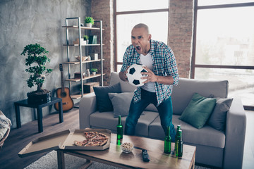 Portrait of delighted funny funky youth hold hand foot ball stand sofa shout yeas triumph victory success wear shirt checked denim jeans apartment indoors house