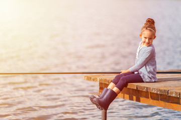 Cute happy little smiling child girl in rubber boots fishing from wooden pier on a lake. Family leisure activity during summer sunny day