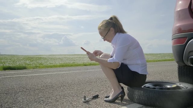 Upset woman crying and calling for help using smartphone. girl sits on spare wheel near car on deserted road.