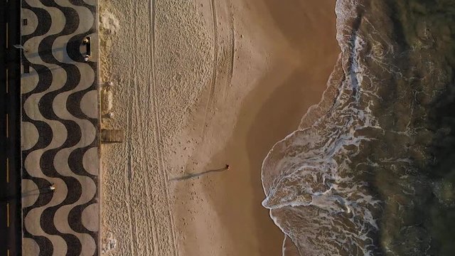 Fixed aerial top down view of Copacabana beach at sunrise with a wave coming in while while a long shadow of a person passes in the sand and on the boulevard pavement. Slowed down from 60fps.