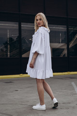 The girl is dressed in a stylish white, lush summer dress, dynamically poses for the photo, posing against street , urban clothing style. Street photography