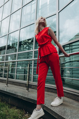 young smiling woman with light hair dressed in a bright red suit with pants and white sneakers on the background of a glass building, urban clothing style. Street photography