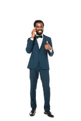 happy african american man in suit talking on smartphone and showing thumb up isolated on white