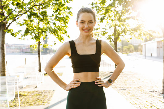 Image of attractive sportswoman smiling and standing with arms on waist during workout in city park