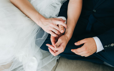 Fototapeta premium Wedding couple sitting on the sofa barefoot. Bride and groom holding hands together. Love story. Elegant clothes. Cosy home interior.