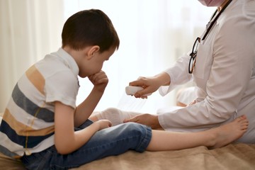 The doctor in a white coat carefully provides first aid. He puts a bandage on the boy's leg. The...