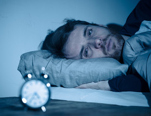 Young man in bed staring at alarm clock trying to sleep feeling stressed and sleepless