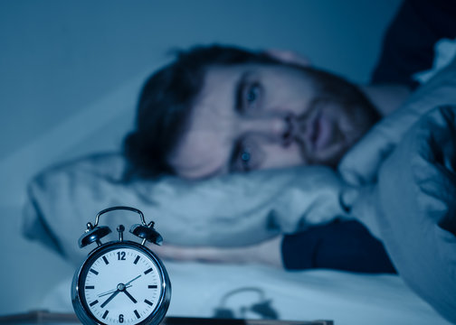 Young man in bed staring at alarm clock trying to sleep feeling stressed and sleepless