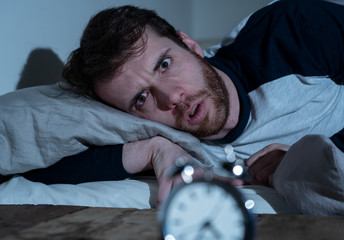 Desperate stressed young man whit insomnia lying in bed staring at alarm clock trying to sleep