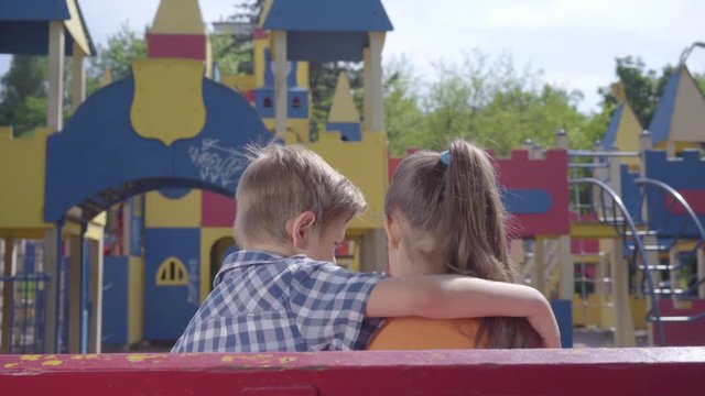 Cute blond boy kissing cheek of a pretty girl sitting on the bench in front of the playground. A couple of happy children. Funny kids in love