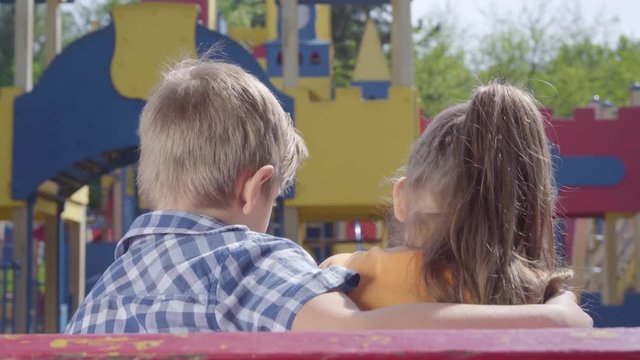 Cute blond boy and a pretty girl sitting on the bench in front of the playground hugging. A couple of happy children. Funny kids in love.