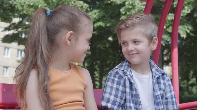 Cute boy and girl sitting on the swing close up in the park, looking at each other with tenderness. A couple of happy children. Funny kids in love