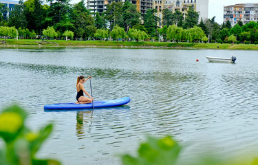 Fototapeta na wymiar Young girl paddling on SUP board on a calm lake at city. Sup surfing woman. Awesome active training in outdoor.