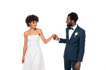 happy african american bridegroom looking at bride while holding hands isolated on white