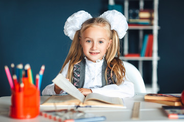 Child and school supplies home Schoolgirl at the table with a textbook and colorful stationery. Girl in school uniform at a Desk in the classroom, holding a textbook, looking at the camera and smiling