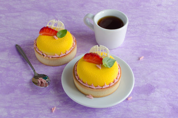Contemporary tropical mini mousse tarts covered with yellow velvet spray decorated with fresh strawberries, basil and chocolate elements with cup of black tee on violet background