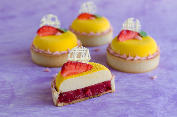 Contemporary tropical mini mousse tarts covered with yellow velvet spray decorated with fresh strawberries, basil and chocolate elements on violet background