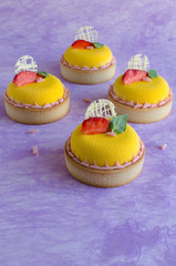 Contemporary tropical mini mousse tarts covered with yellow velvet spray decorated with fresh strawberries, basil and chocolate elements on violet background