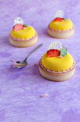 Contemporary tropical mini mousse tarts covered with yellow velvet spray decorated with fresh strawberries, basil and chocolate elements on violet background