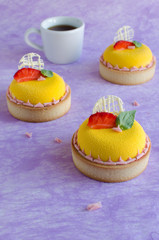 Contemporary tropical mini mousse tarts covered with yellow velvet spray decorated with fresh strawberries, basil and chocolate elements on violet background