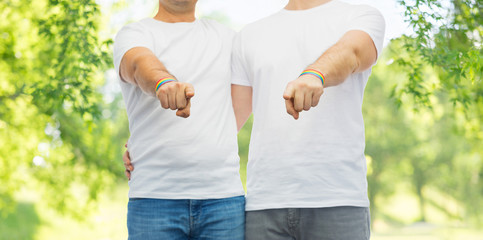 lgbt, same-sex relationships and homosexual concept - close up of hugging male couple wearing gay pride rainbow awareness wristbands and pointing fingers at you over green natural background