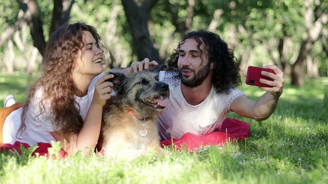 A young cheerful couple taking a selfie at the park with their shaggy dog on the summer day. Having a good time with a family.
