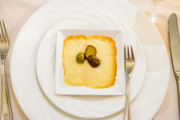 grilled melted gruyere cheese appetiser topped with black and greeen olives on a square plate