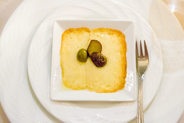 grilled melted gruyere cheese appetiser topped with black and greeen olives on a square plate