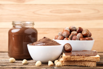Bread with melted chocolate, glass jar and hazelnuts on brown wooden table
