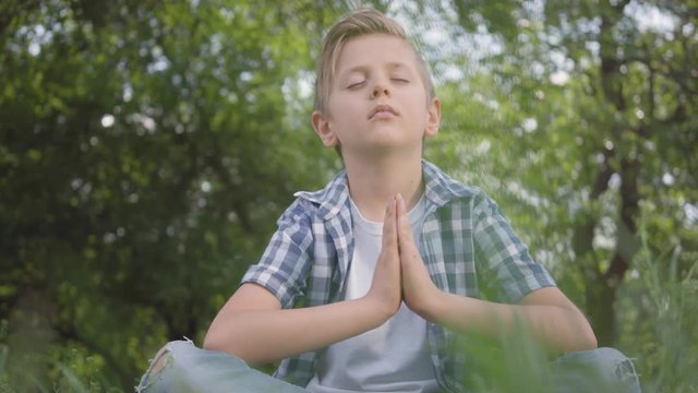Cute little handsome boy sitting on the grass meditating. Child practices yoga. The spiritual development of kids. Summertime leisure.