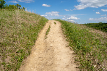 Dirt hiking trail surrounded by green grass goes up a hill on a sunny day, Leading lines