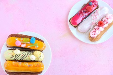 Tasty French eclairs with icing, cream, fresh berries and sugar decor elements. Set of delicious eclairs with creative colorful decor on white plates on the pink textured table. Homemade profiteroles 