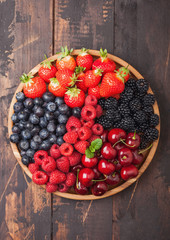 Fresh organic summer berries mix in round wooden tray on dark wooden table background. Raspberries, strawberries, blueberries, blackberries and cherries.
