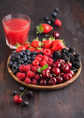 Fresh organic summer berries mix in round wooden tray with glass of juice on dark wooden table background. Raspberries, strawberries, blueberries, blackberries and cherries.