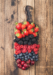 Fresh organic summer berries mix on vintage wooden chopping board on light wooden table background. Raspberries, strawberries, blueberries, blackberries and cherries.