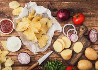 Fresh organic homemade potato crisps chips with sour cream and red onions and spices on wooden background. With fresh yellow potatoes on chopping board.