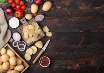 Fresh organic homemade potato crisps chips with sour cream and red onions on chopping board on dark wooden background. Tomatoes with green onion and bowl of ketchup.