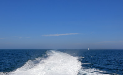 Boat wake in the sea on a brilliant sunny day with distant sailboat.