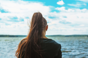 Fototapeta na wymiar Young woman with long brown ponytail hair stands on the coast and looks into the distance. Silhouette of a girl view from the back on the background of the sea horizon